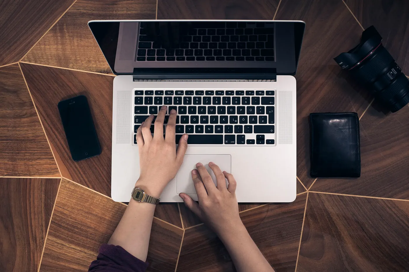 Illustrative handover: hands on a laptop keyboard, seen from above, with a phone and a wallet on a wooden desk